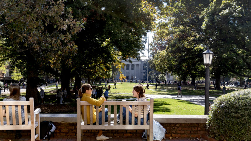 Two people talking to each other on a bench facing Polk Place at UNC Chapel Hill